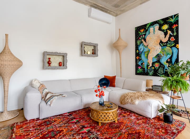 Un salon chaleureux avec un canapé blanc en forme de L, un tapis aux motifs colorés et une décoration éclectique, avec une peinture représentant des personnages entrelacés dans une végétation luxuriante.