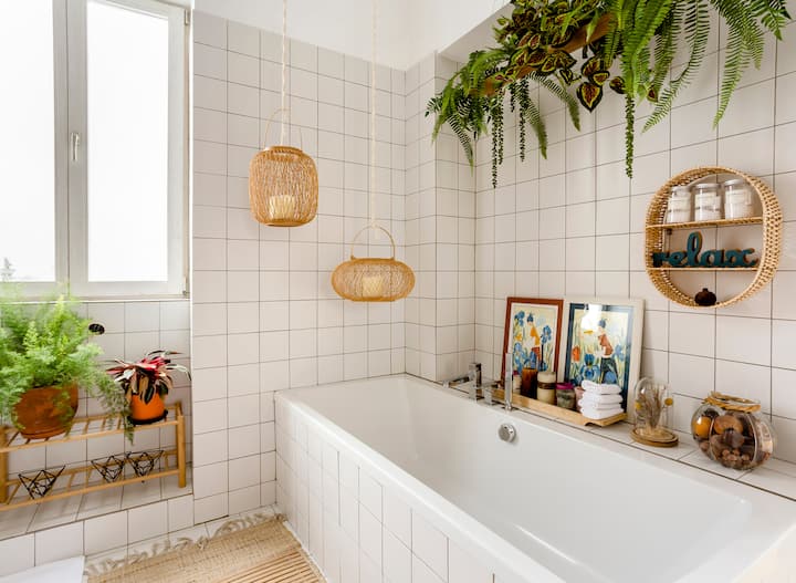 Une salle de bain chaleureuse avec un mur en carrelage blanc, une baignoire rectangulaire, des suspensions en osier, des plantes en pot, des cadres, ainsi qu'une étagère avec des bougies et une décoration représentant le mot « relax ».