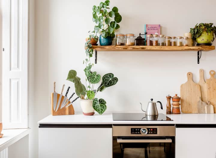 Une cuisine lumineuse et moderne avec un plan de travail blanc élégant, une cuisinière intégrée, une bouilloire, un set de couteaux et une plante en pot. Au-dessus du plan de travail est fixée une étagère en bois sur laquelle sont posés des pots, un livre coloré et d'autres plantes.
