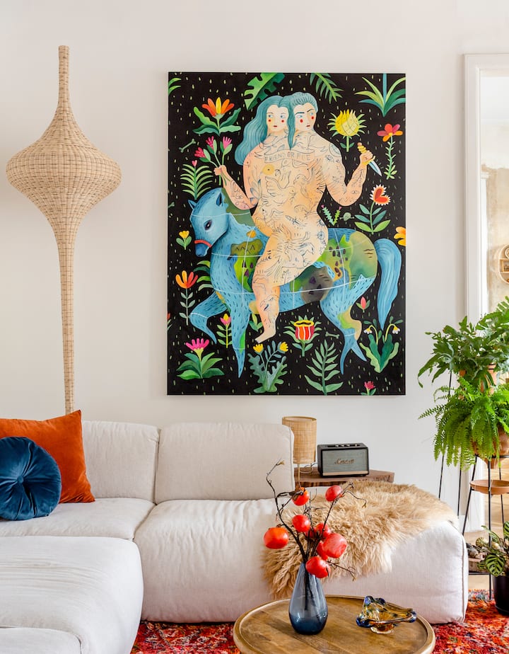 Un salon à l'ambiance chaleureuse et artistique, avec un canapé blanc, une lampe en rotin et des plantes en pot, ainsi qu'un tableau coloré accroché sur le mur du fond et représentant deux personnages enlacés sur un cheval bleu entouré de fleurs.