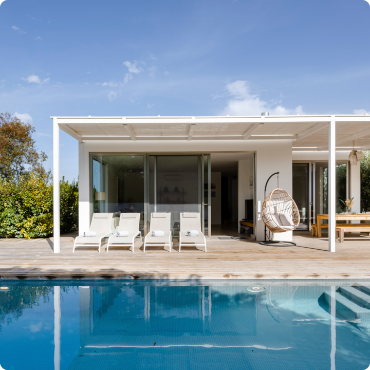 Une terrasse couverte moderne donnant sur une piscine, avec quatre chaises longues blanches, une chaise en rotin suspendue et un coin repas sous une pergola, le tout sous un ciel bleu vif et avec de la végétation à l'arrière-plan.