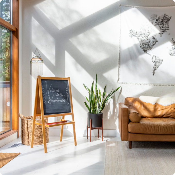 Un salon lumineux avec un canapé en cuir, une plante en pot et un tableau noir où est écrit « Welcome to the Woodlands » et derrière lequel est posé un panier tissé. Sur le mur du fond, une tapisserie murale représente une carte du monde.