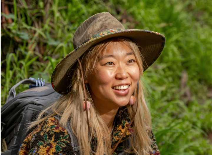 Une personne vêtue d'un pull coloré et d'un chapeau à large bord avec un motif floral porte un sac à dos de randonnée. Elle est entourée d'une végétation luxuriante lors d'une aventure en plein air.