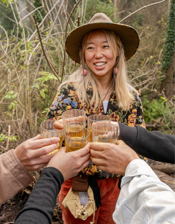 Quatre mains tenant des verres de boisson légère, trinquant ensemble dans un cadre extérieur entouré de verdure et d'arbres, avec une hôte portant une veste à fleurs et un chapeau à large bord en arrière-plan.