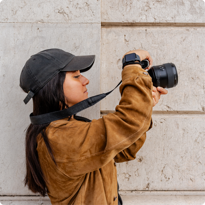 Une personne portant une casquette noire et une veste marron tient un appareil photo professionnel et prend une photo devant un mur de pierre texturé.