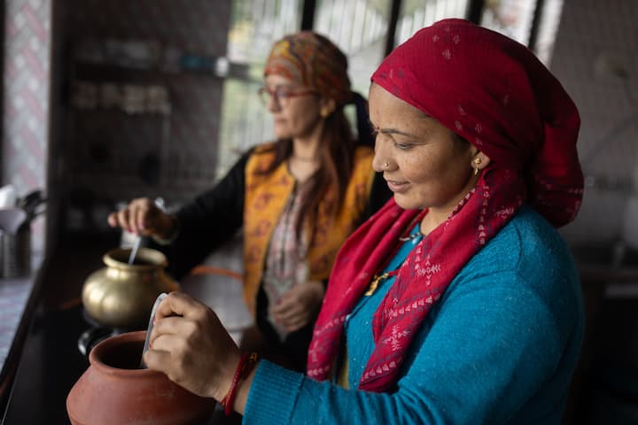 Two women in a rural kitchen setting preparing food, with one stirring a pot and the other handling a traditional clay vessel, highlighting local entrepreneurship and homestay hosting initiatives.