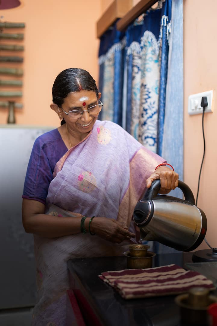 An Indian woman wearing a pastel saree pours tea from an electric kettle into a brass cup in a cozy kitchen with blue patterned curtains.
