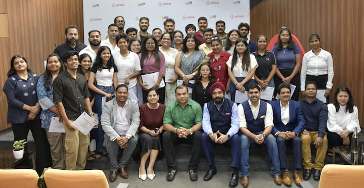 A group of individuals posing together in a formal setting, holding certificates, with an Airbnb and partner organization-branded backdrop behind them.