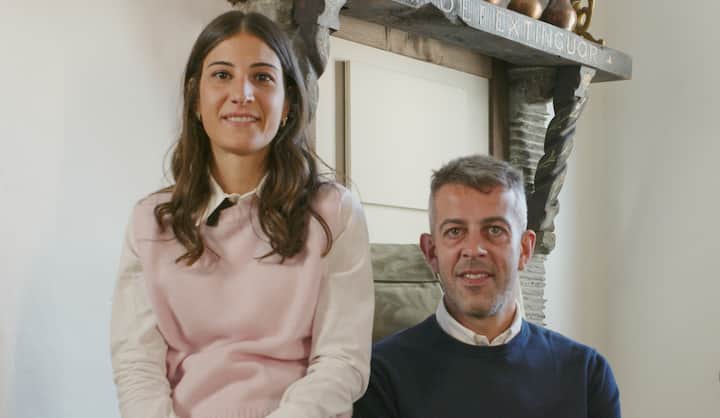 Two people seated in a warmly lit living room with a rustic fireplace in the background, featuring wooden beams and decorative details.