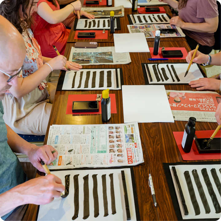 Des personnes participant à un atelier de calligraphie japonaise tracent des traits d'encre noire sur du papier, entourées d'outils et de matériel tels que des bouteilles d'encre et des journaux.