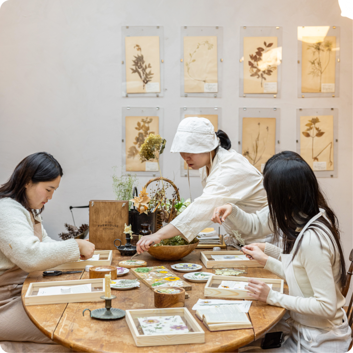 Dans une pièce décorée de fleurs séchées et d'impressions botaniques, des personnes participant à un atelier créatif arrangent des fleurs pressées dans des cadres.