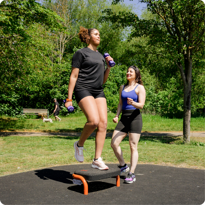 Une personne monte sur un stepper avec des haltères à la main, guidée par une autre personne dans un parc entouré d'arbres.