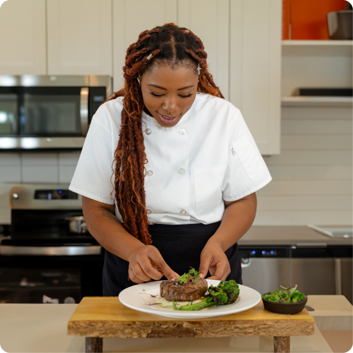 Dans une cuisine moderne, une cheffe vêtue d'une tenue blanche dresse un steak accompagné de brocoli et de jeunes pousses.