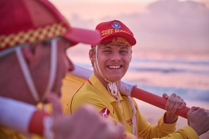 Two lifeguards in yellow uniforms and red caps labeled 'Surf Rescue' holding a rescue paddle on a beach during sunset, with waves in the background.