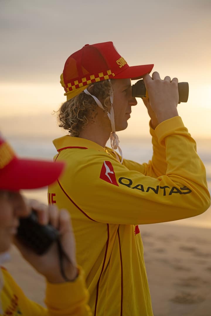 A lifeguard in a yellow and red uniform holds binoculars while observing the beach during sunset.