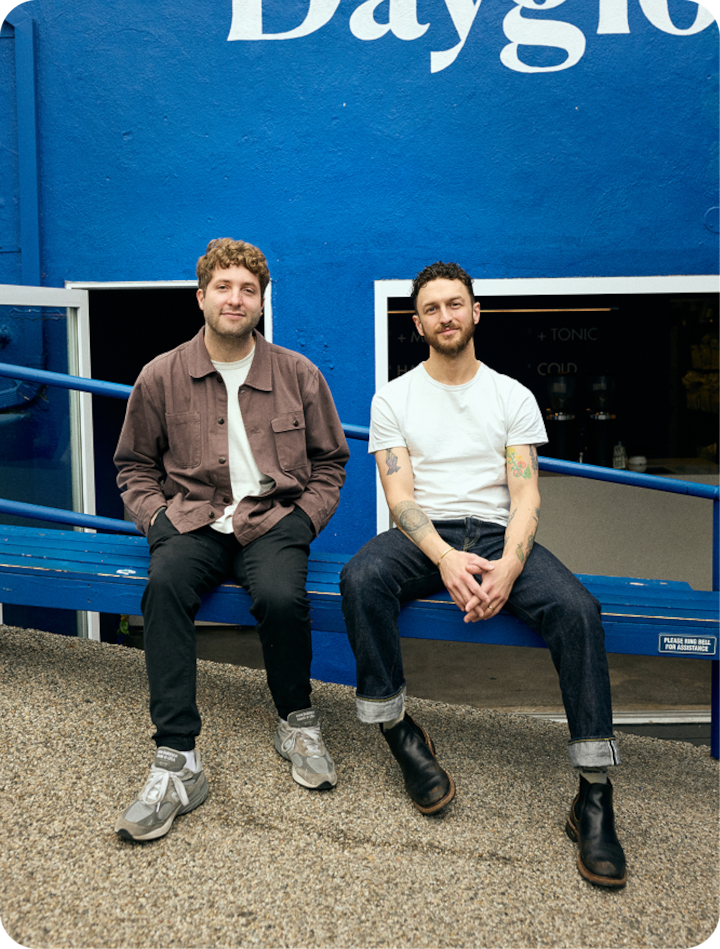 Deux personnes sont assises sur un banc bleu à l'extérieur. Derrière elles, un mur bleu vif porte l'inscription « Dayglow ». Le cadre est composé d'éléments simples et épurés.