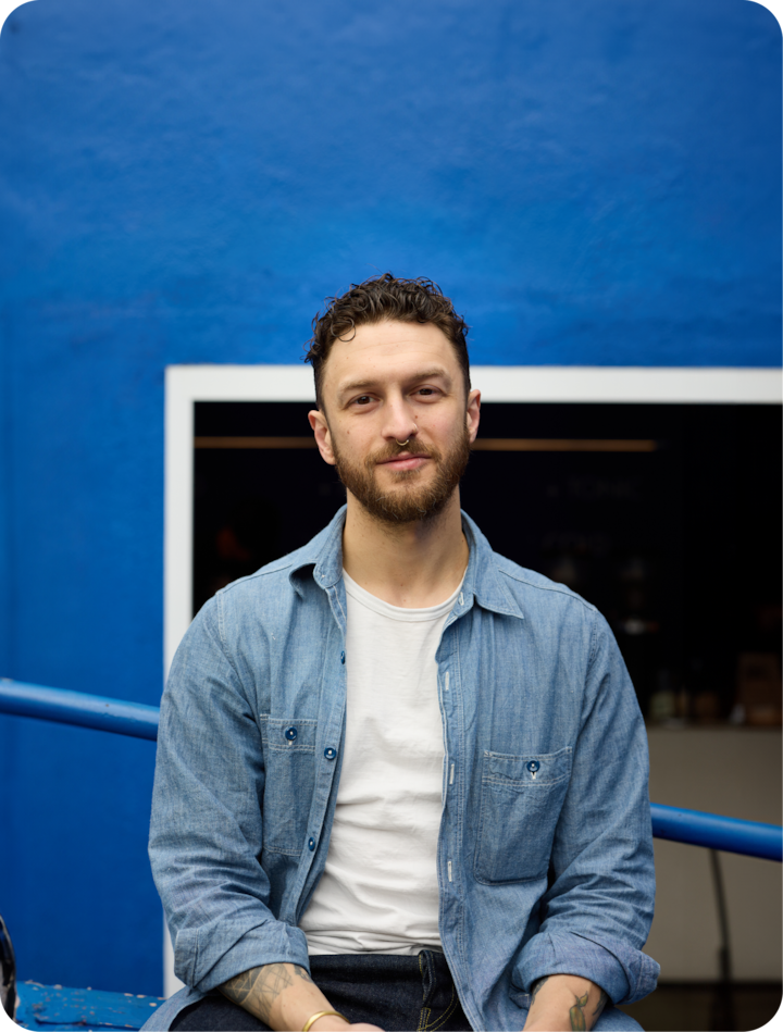 Une personne est assise dans une posture détendue, avec un arrière-plan sobre et épuré : un mur bleu vif, avec un aperçu discret d'un espace intérieur visible à travers une porte.