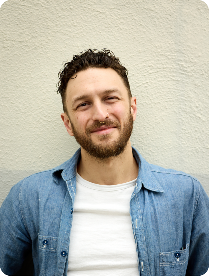 Photo du haut du corps d'une personne qui se tient devant un mur clair, neutre et minimaliste. Elle porte un t-shirt blanc et une chemise en jean bleue déboutonnée.