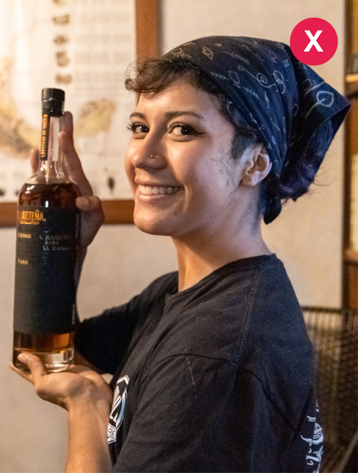 Une personne tenant une bouteille de liqueur artisanale avec une étiquette sombre se tient devant un mur avec une affiche de cartes illustrées.