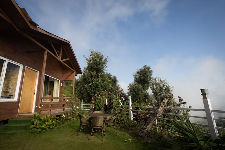 A cozy wooden house with a balcony surrounded by a lush garden, outdoor seating, and overlooking a misty landscape in Uttarakhand.
