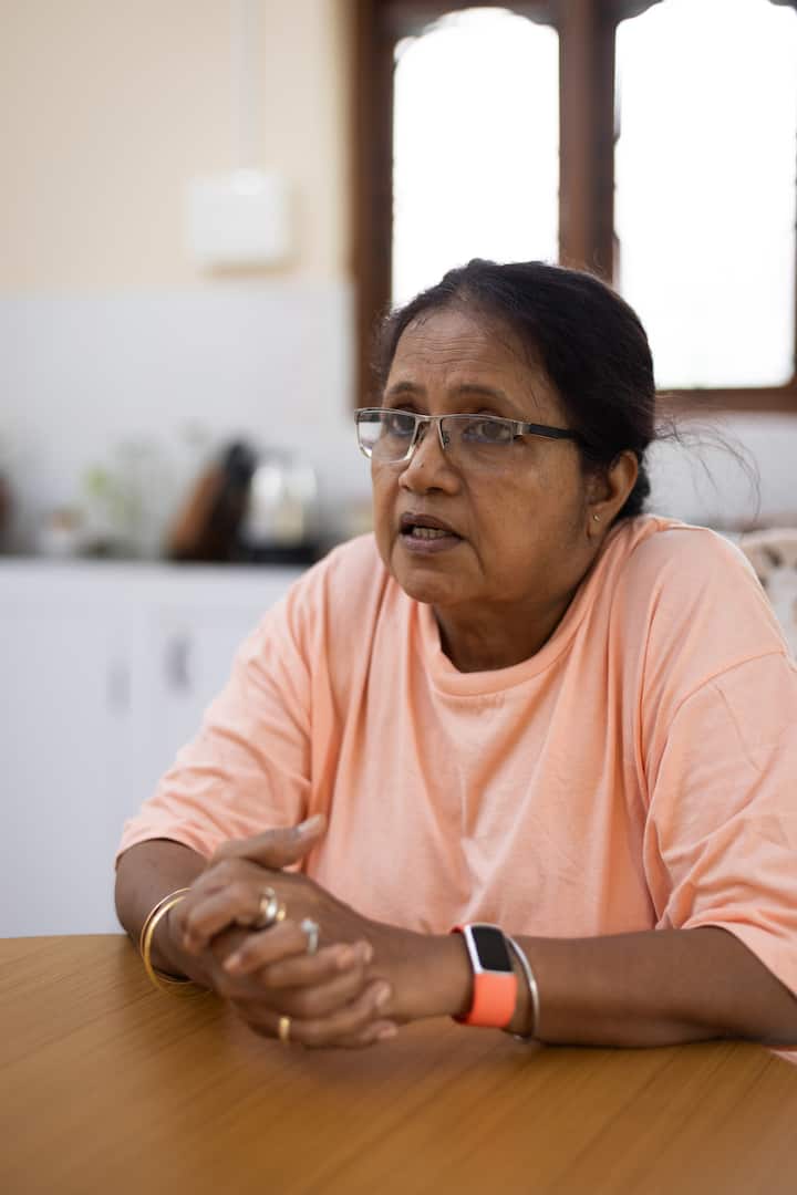 A person wearing an orange shirt, gold jewelry, and an orange smartwatch sits at a wooden table in a bright kitchen with white cabinets and a window in the background.