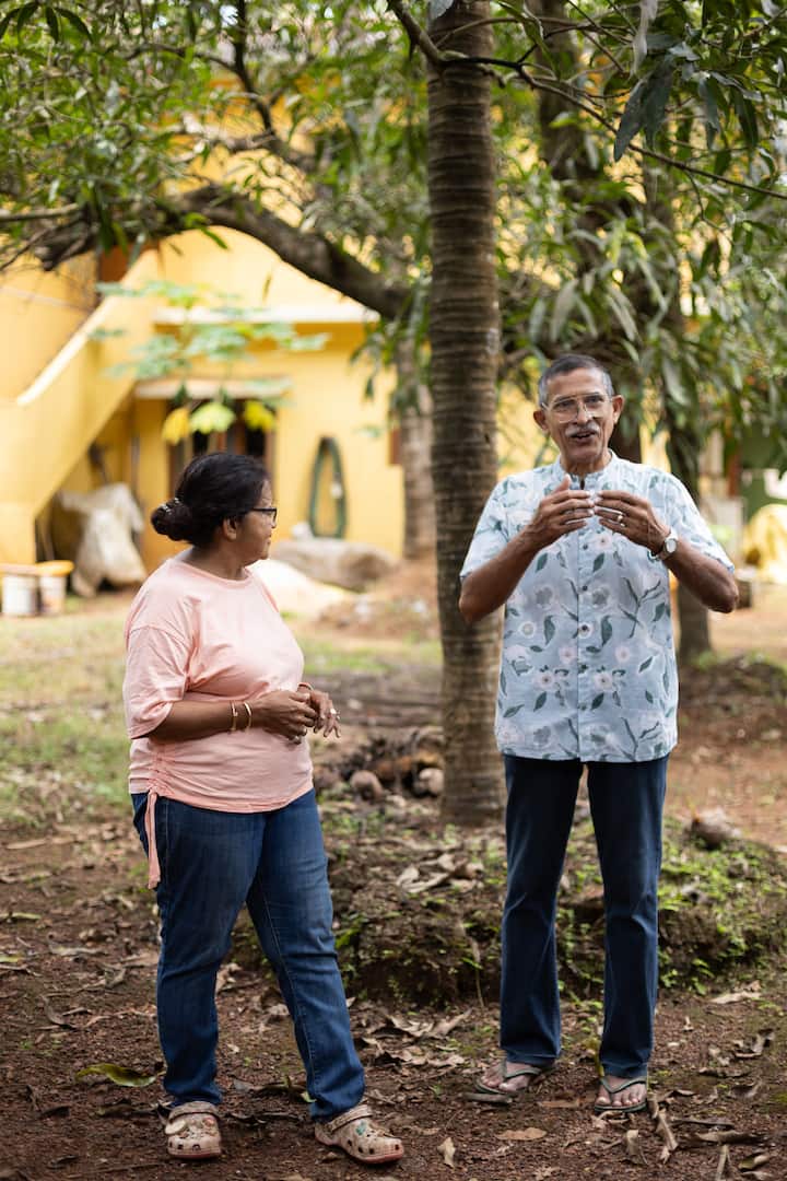 A man and woman standing outdoors in a garden with a yellow house and lush greenery in the background, talking under a tree.