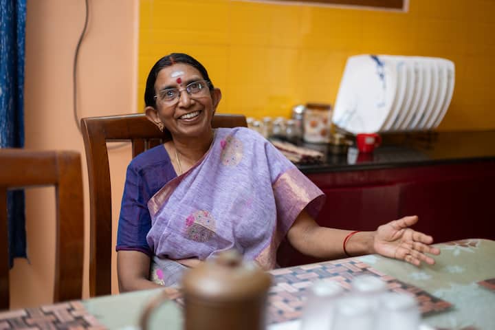 A woman wearing a purple sari sits at a dining table in a warmly lit kitchen, with crockery and utensils visible in the background.