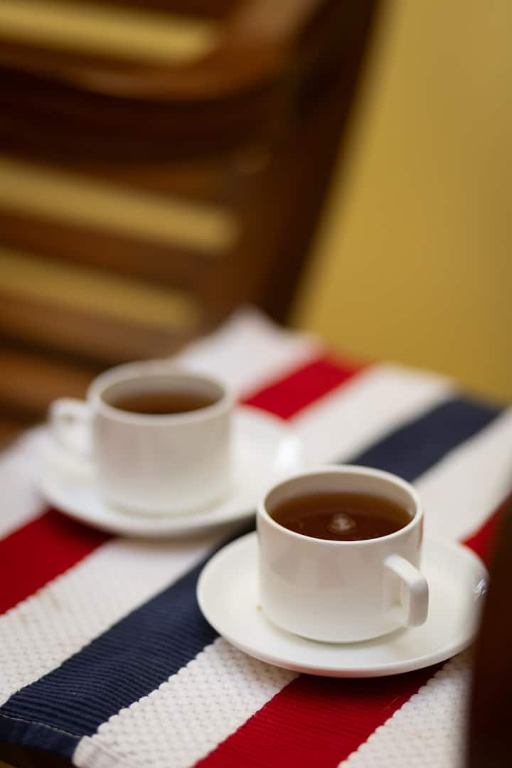 Two white teacups filled with hot tea placed on a red, white, and blue striped tablecloth, with blurred wooden chair in the background.