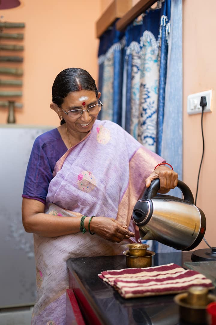 A woman in Tamil Nadu wearing a lavender saree pours hot water from an electric kettle into a traditional brass cup on a kitchen counter with blue curtains in the background.