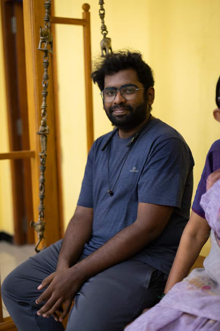 A man wearing a navy blue t-shirt and gray pants sits on an ornate wooden swing with brass decorations in a brightly lit room. Part of a woman in a purple sari is visible beside him.