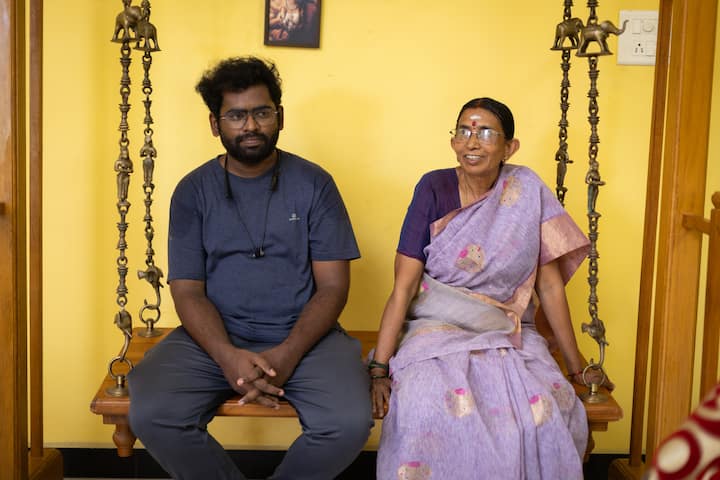 A cozy indoor space featuring two traditional wooden swing seats with ornate metal chains, a man in a casual shirt, and a woman wearing a light purple saree, against a yellow wall with traditional decor.