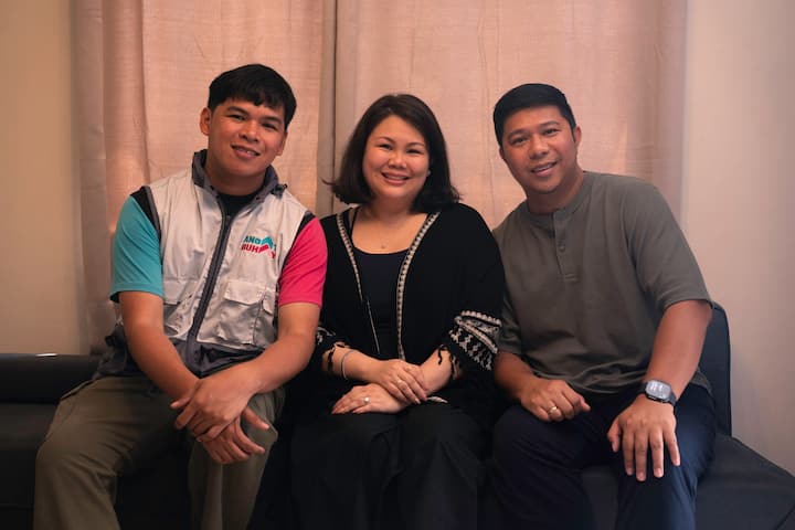 Airbnb.org-gjesten Menard med Airbnb.org-vertene Mia S. og Pau G. sitter på en sofa sammen og smiler til kameraet.