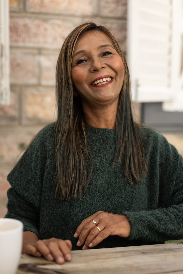 Person wearing a dark green sweater seated at a wooden table with a cup of coffee in the foreground, stone wall and white shutters visible in the background.