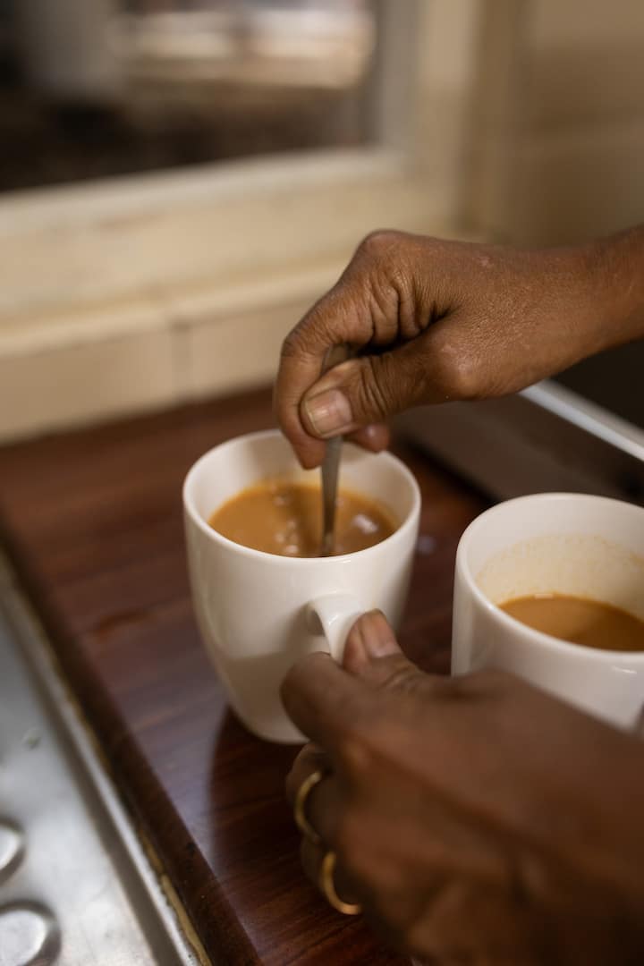 Close-up of a hand stirring tea in a white mug, with another mug of tea beside it on a wooden countertop.