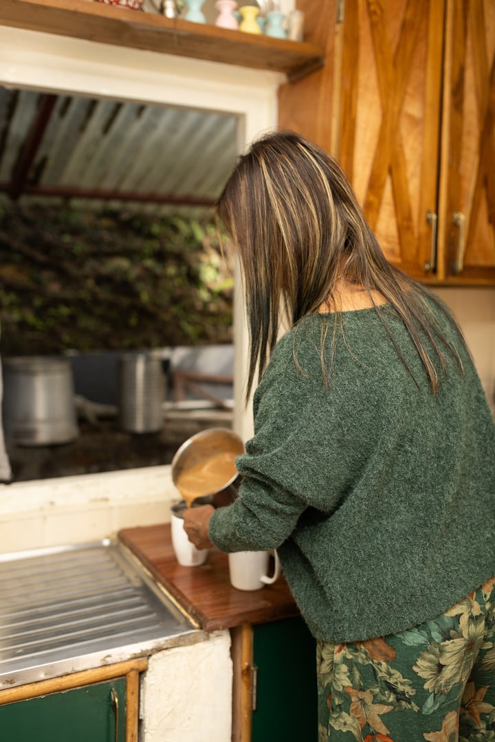 Anne, a host from Meghalaya, pours tea into a mug in a cozy kitchen with wooden cabinets and a window overlooking greenery.
