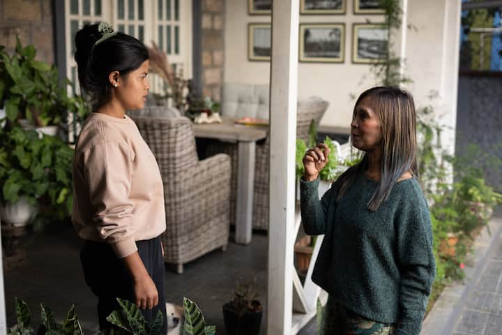 Two women standing on the porch of a homestay surrounded by greenery, engaged in a conversation, with wicker furniture and framed photos visible in the background.