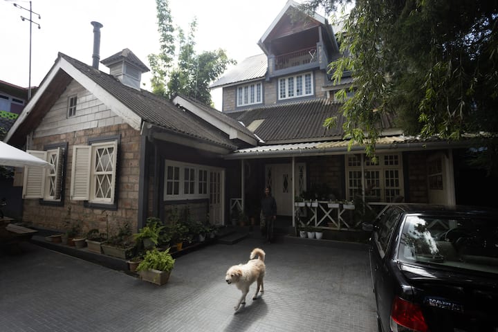 A charming stone house with white shutters surrounded by greenery in Meghalaya, a dog walking in the driveway and a person standing near the entrance.
