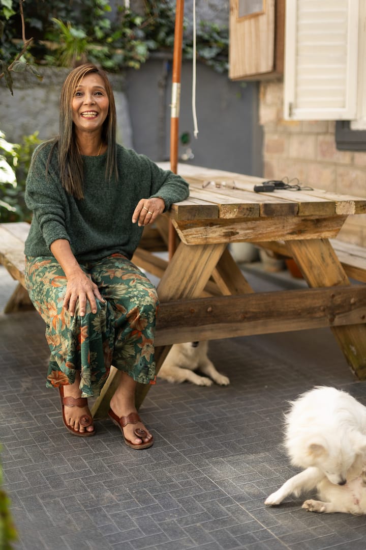 A person sitting at a wooden picnic table in a serene outdoor setting with greenery in the background, wearing a green sweater, a floral-patterned skirt, and brown sandals, with a white dog lying on the tiled floor nearby.