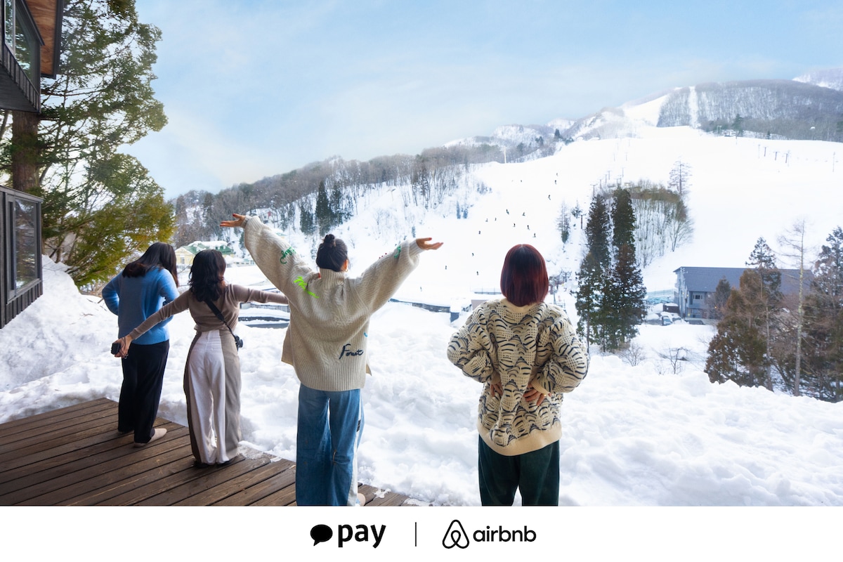 스키 리조트와 산을 배경으로 눈 덮인 발코니에 네 사람이 서 있습니다. 한국에서 진행하는 에어비앤비와 카카오페이 겨울 휴가 홍보 이미지입니다.