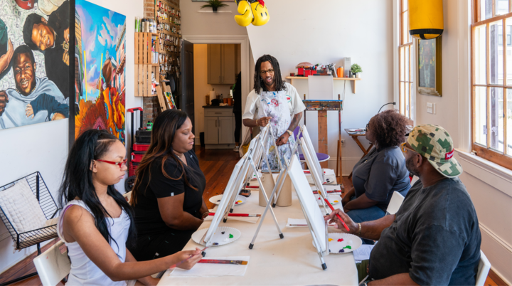 Un groupe de personnes assises à une longue table dans un studio d'art bien éclairé participent à un atelier de peinture. Chacune travaille sur sa propre toile avec des palettes de peinture et des pinceaux, entourée d'œuvres d'art colorées et d'un décor créatif.