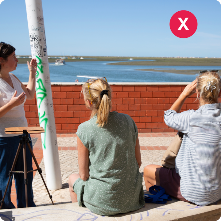 Une scène en plein air d'un atelier au bord de l'eau ; deux femmes sur un banc discutent avec une personne qui fait des gestes à côté d'un poteau couvert de graffitis.