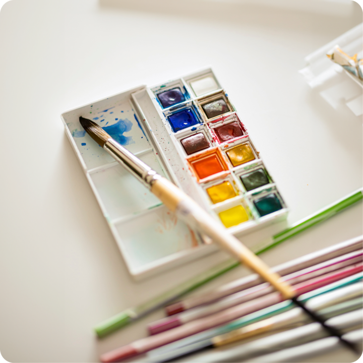 L'espace de travail d'un artiste avec un ensemble d'aquarelles et plusieurs pinceaux, l'un d'entre eux ayant de la peinture bleue sur sa pointe. L'accent est mis sur la créativité et les expériences artistiques locales.