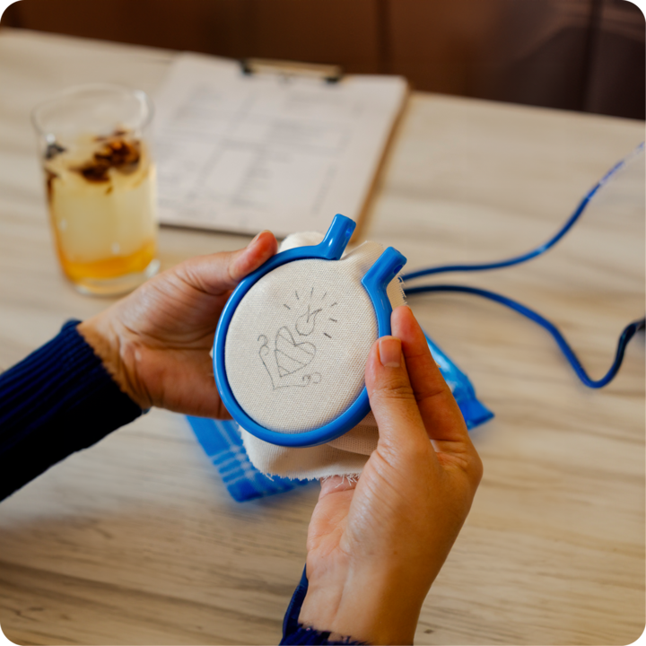 Plan rapproché d'une personne tenant un cerceau de broderie bleu avec un motif de cœur et d'aiguille, capturant l'esprit créatif des expériences d'atelier pratique.