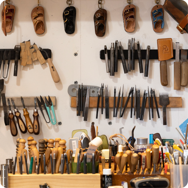 Un mur d'atelier présentant un éventail d'outils pour travailler le cuir, y compris des poinçons, des alènes et du matériel de sculpture, avec des petites sandales en cuir suspendues au-dessus.