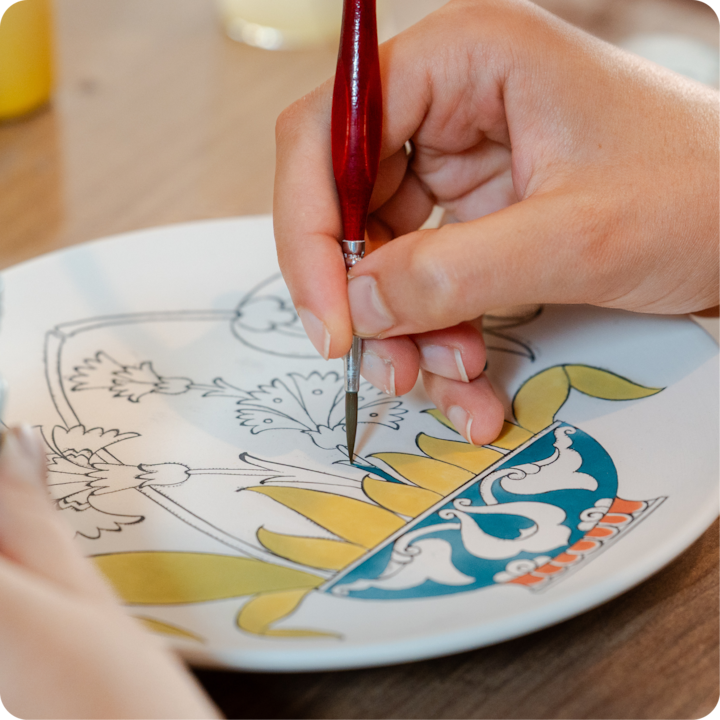 Lors d'un atelier culturel local, une personne peint soigneusement des détails jaunes, bleus et verts sur une assiette décorative en céramique.