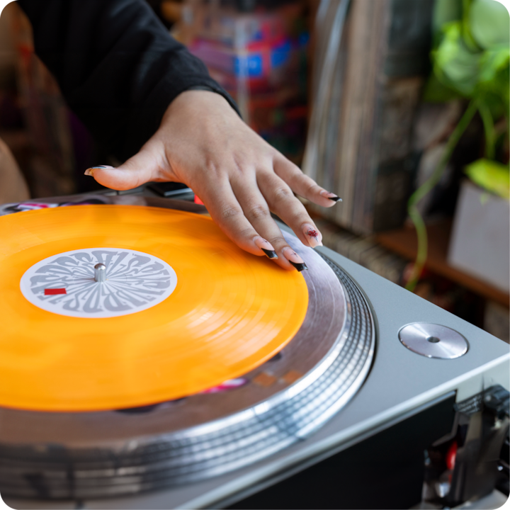 Un disque vinyle orange tourne sur une platine sous la main d'une personne, avec un atelier coloré et créatif en arrière-plan.
