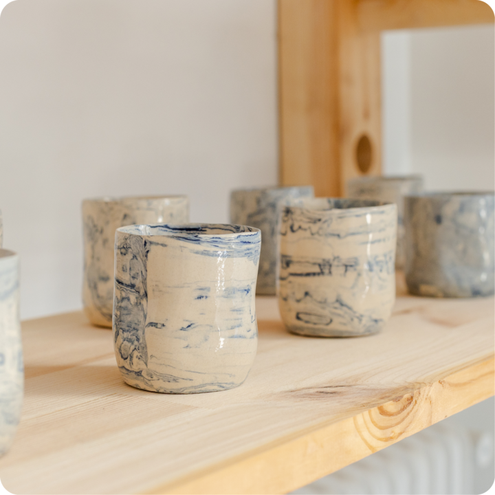 Gros plan sur des tasses en céramique artisanales ornées de tourbillons bleus et beiges, disposées sur une étagère en bois clair dans un atelier créatif.