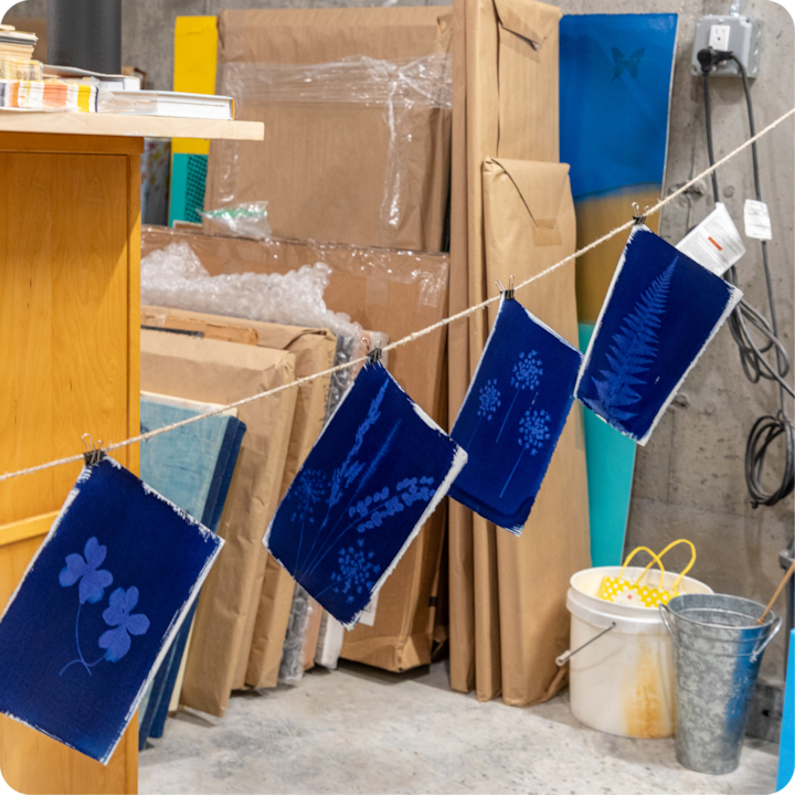 Quatre impressions cyanotype bleues aux motifs botaniques sont suspendues à une ficelle dans un atelier entouré de matériel d'art et d'emballage.