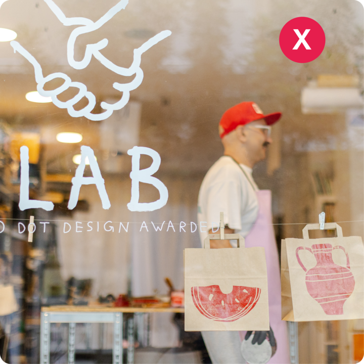 Des sacs en papier portant des motifs rouges estampés représentant une tranche de pastèque et un vase suspendu à une ficelle devant une vitrine portant le texte « LAB », montrant un environnement d'atelier créatif à l'intérieur.