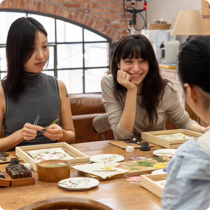 Un atelier créatif pratique avec des participants qui conçoivent des objets artisanaux en fleurs pressées sur une table en bois, avec un éclairage naturel provenant de grandes fenêtres et un arrière-plan de murs en briques apparentes et d'outils d'atelier.